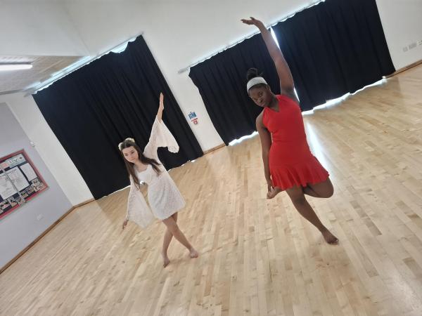 Year 11 Dance Exam Photo of two dancers mid performance in the dance studio. One dancer is wearing a white dress, the other a red dress, they both have their legs crossed and their left arm in the air.