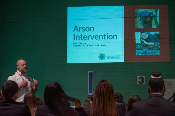 Photograph of Joe Gacon, the Arson Intervention Lead at Cambridgeshire fire and rescue. He is stood in front of student delivering his talk with a slide projected behind him which reads Arson Intervention