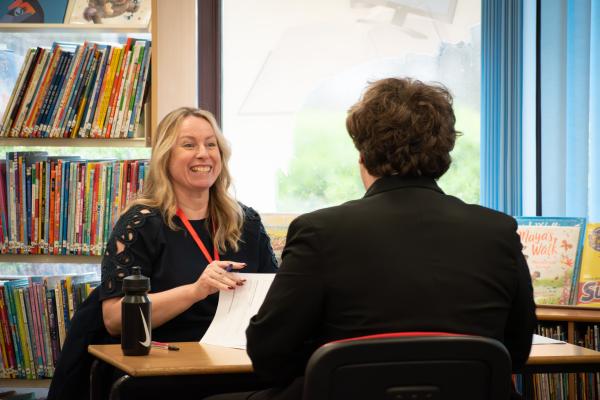 Photo of a student and an interviewer sat at a desk in a library.  The female interviewer is laughing, the student has his back to the camera.