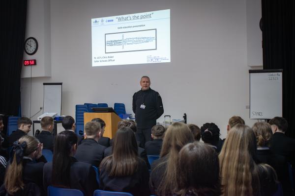 Photo of PC Chris Baker stood in front of an audience of Year 9 students.  There is a projection in the background detailing the topic of the talk - "What's the Point" and a picture of a knife made out or word art.