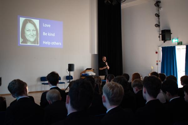 Photo of Anthony stood at the front of our Assembly Hall in front of our Year 9 students who are sat in chairs watching.  Behind him is a power point presentation showing a photo of his mum and the words Love, Be Kind, Help Others